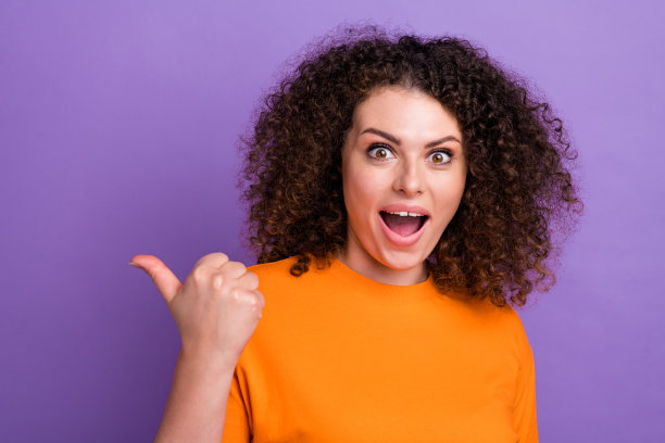 印象深刻的时髦女士穿着橙色t恤张开嘴指着拇指空白空间孤立的紫色背景图片下载