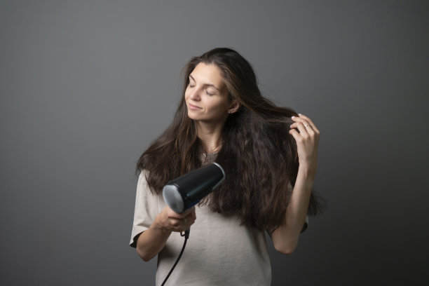 照片中，一名年轻女子心满意足地用吹风机在中性背景下梳理自己飘逸的棕色头发，捕捉了个人打扮的瞬间。图片下载
