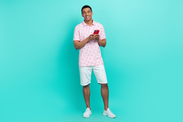 智能男子的全身照片，穿着印花t恤白色短裤，拿着智能手机阅读电子邮件，孤立在绿松石色的背景图片下载