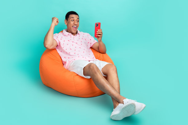 混血的全身照片有魅力的男人穿马球躺在扶手椅上看智能手机赢赌注孤立的绿松石色背景图片下载