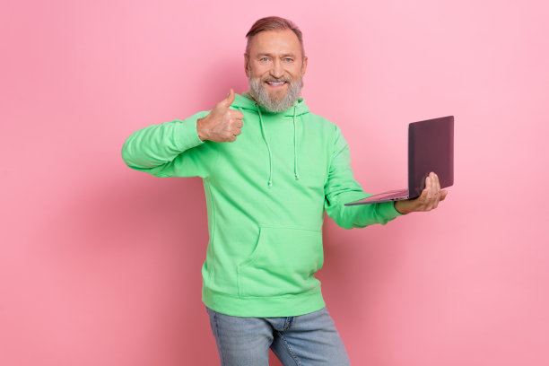 照片的积极自信的家伙穿着绿色卫衣显示拇指向上工作的现代设备孤立的粉红色背景图片下载