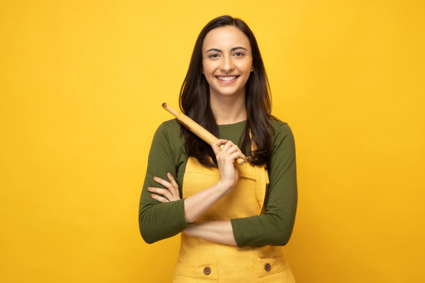 印度女厨师戴着围裙，拿着木制擀面杖，孤立地站在黄色背景照片上图片下载