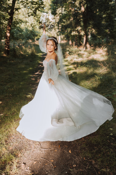 一位美丽的新娘身穿白色长裙，戴着长长的面纱，手持花束在公园里盘旋。后面的照片。年轻女子，艺术照片，婚礼，新娘，开放的肩膀。光。图片下载