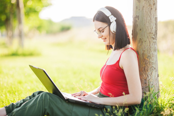 深棕色头发的女孩戴着白色耳机在公园的树下使用笔记本电脑图片下载