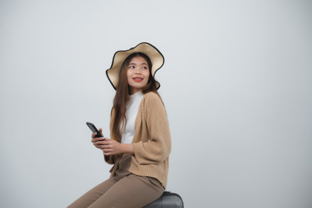 亚洲微笑的女游客带着牙套，戴着帽子，坐着的行李拿着手机，复制空间去旅行假期孤立的蓝色工作室背景。旅行之旅的概念。图片下载