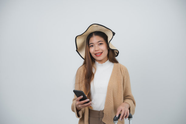 亚洲微笑的女游客带着牙套，戴着帽子，拿着行李，拿着手机，复制空间去旅行假期孤立的蓝色工作室背景。旅行之旅的概念。图片下载