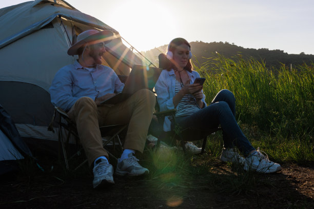白人男性在笔记本电脑上工作，女性通过耳机用智能手机听音乐，在帐篷外的阳光背景下，图片下载