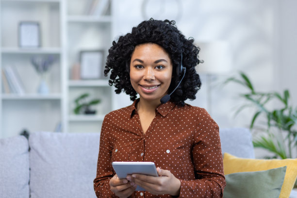 一个年轻的非洲裔美国女人微笑着坐在家里的沙发上，戴着耳机，用着平板电脑。特写照片图片下载
