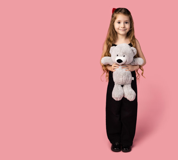 一个小女孩的全身画像，穿着黑色的牛仔布，怀里抱着一只灰色的泰迪熊，开心地看着镜头，献身于对玩具的热爱。浅粉色背景图片。图片下载