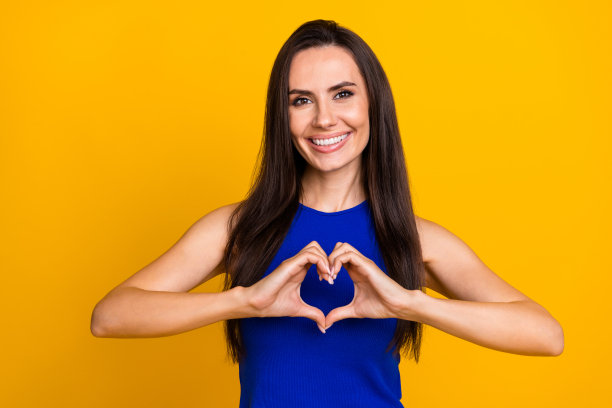 照片中，兴奋可爱的女人穿着蓝色上衣，露出手臂，心形手势，孤立的黄色背景图片下载