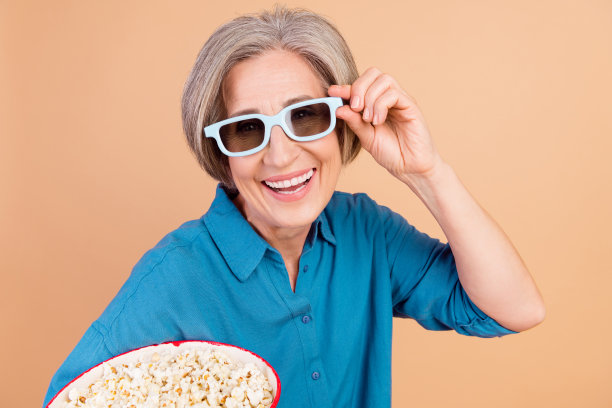 照片中，长着牙齿、笑容灿烂的女人留着波波头，穿着蓝色衬衫，触摸3d眼镜，在电影院里拿着爆米花，背景是柔和的颜色图片下载