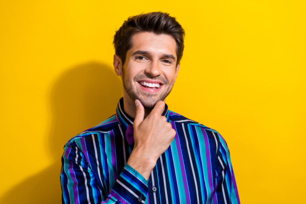 一个穿着条纹衬衫、笑容满面的男人，手放在下巴上，在鲜艳的黄色背景上思考着商业理念图片下载