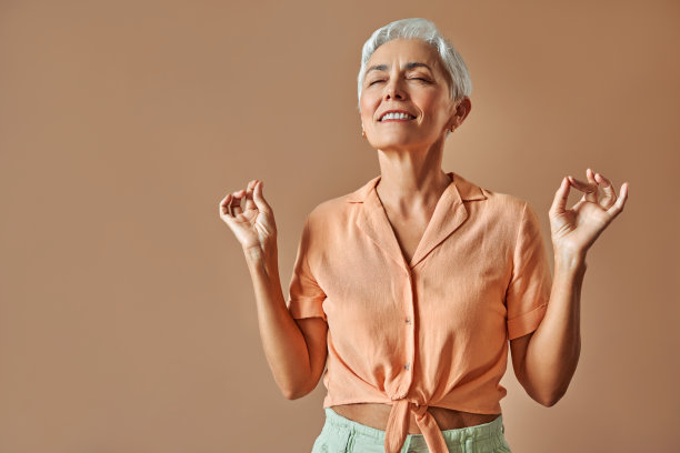 美丽的现代微笑真诚时尚成熟的白发短发女人在柔和的橙色t恤表达平静的情绪和闭着眼睛沉思，站在米色背景。图片下载