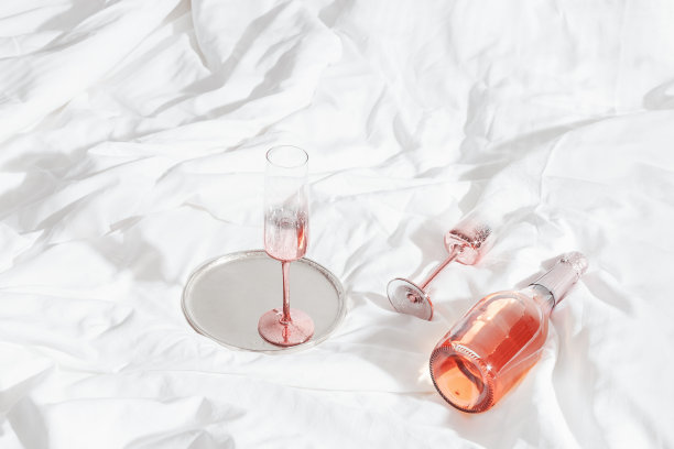 粉红色的香槟酒杯与金属光泽和一瓶玫瑰起泡酒在白色的床单。生活唯美的照片，星光滤光。情人节佳节，爱情观念，浪漫相遇。图片下载