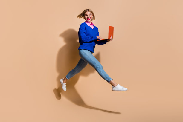 跳跃的年轻商业女性自由创业与笔记本电脑共同工作孤立在米色背景的全长照片图片下载