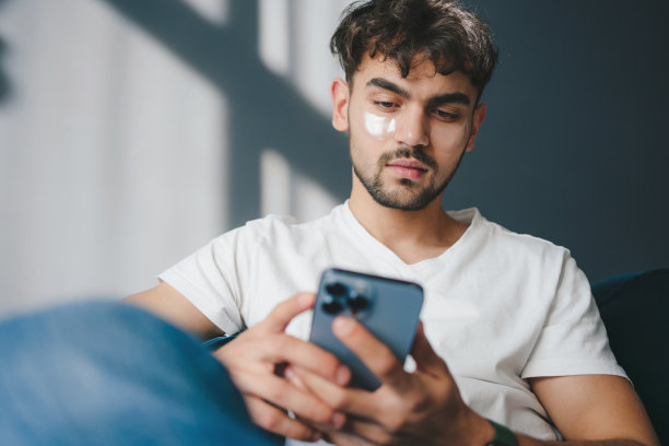 一个穿着休闲服、面带微笑的帅哥坐在客厅沙发上玩手机的照片。现代的生活方式。互联网技术。在线交流。房子内部。手机应用程序。图片下载