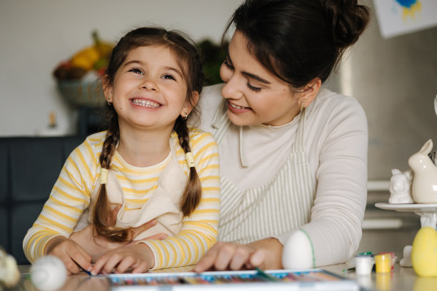 可爱的小女孩和妈妈在厨房的照片。美丽的女人帮助她的女儿画复活节彩蛋图片下载
