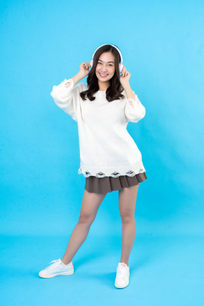年轻的亚洲女模特全身形象，长发，微笑，眼神交流，穿着白大褂，用手触摸耳机。在摄影棚里，蓝色的背景图片下载