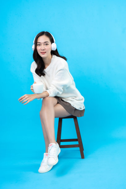 亚洲长发女模特坐下来放松，通过app用无线耳机听音乐，端着一杯咖啡。坐在椅子上，在蓝色背景的摄影棚里拍照图片下载