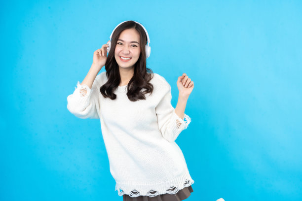 年轻的亚洲女模特的半身像长头发在工作室，蓝色背景。用你的手握住耳机。穿着白大褂听着音乐，开心地笑着图片下载