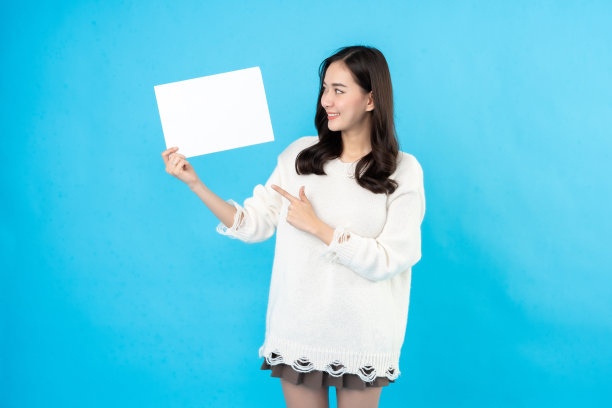 长头发的亚洲女模特指着你手里的白纸。为文本或符号词留出空间。在蓝色背景的摄影棚里拍照，穿白大褂图片下载