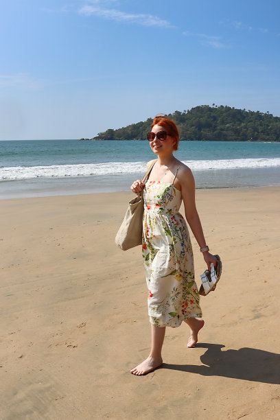 特写照片中，红头发的女人穿着太阳裙，戴着太阳镜，赤脚走在海边潮湿的沙滩上，背着一个肩袋，手里拿着凉鞋，阳光明媚，晴朗的蓝天，前景的焦点图片下载