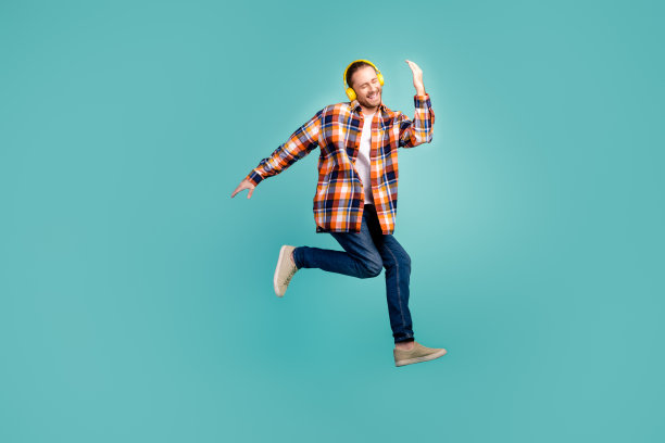 穿着格子衬衫在空中跳舞的滑稽男子的全尺寸照片，在蓝绿色背景上用蓝牙耳机听音乐图片下载