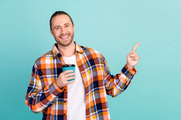 照片中，长着胡子和长发、笑容满面的男子手持一杯咖啡，在蓝绿色的背景上指向空的空间图片下载