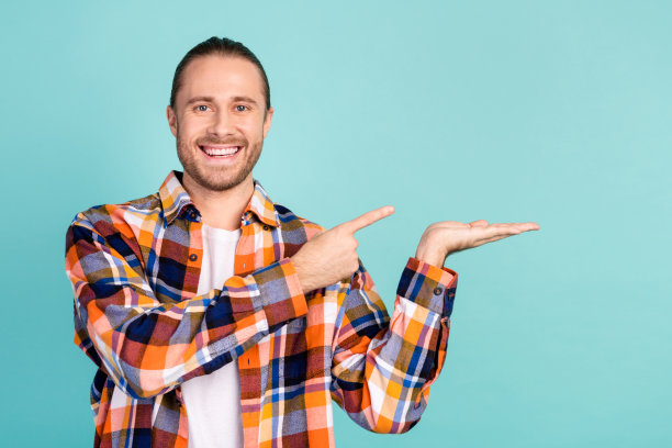 照片中的酷男人留着长发，穿着格子衬衫，指向手掌上的物体，在蓝绿色背景上孤立的空白空间图片下载