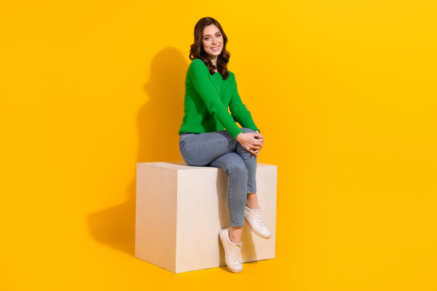 穿着绿色套头衫和牛仔裤的开朗女士全身照片，坐在讲台上可爱的模特在黄色背景上孤立等待图片下载