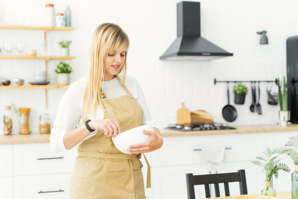 一个欧洲女孩，系着米色围裙，站在白色的厨房里，用手打蛋器在白色盘子里搅打奶油图片下载