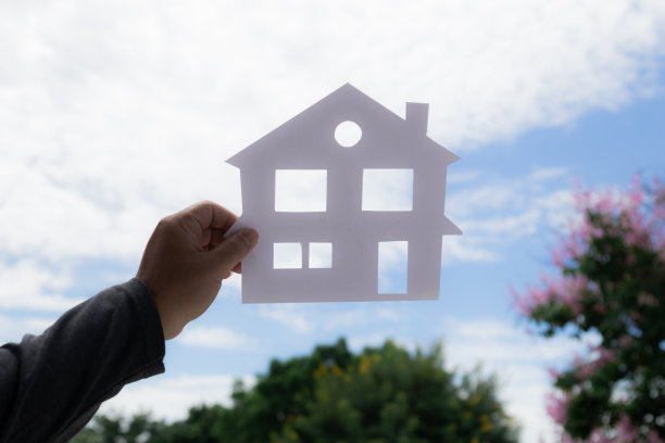 女人手持剪纸的房子图片在美丽的蓝天背景下，住房贷款的生活方式的概念，人们的梦想或稳定的生活，想要有一个房子，富裕的生活规划图片下载