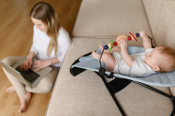 一位年轻的母亲在笔记本电脑上远程工作，一个2.5个月大的婴儿坐在摇椅上玩玩具，关注他。图片下载