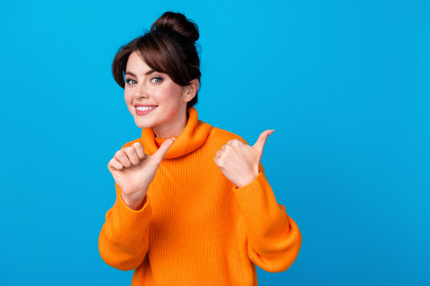 照片中开朗积极的女人穿着橙色毛衣，露出两个拇指，背部空白，孤立的蓝色背景图片下载