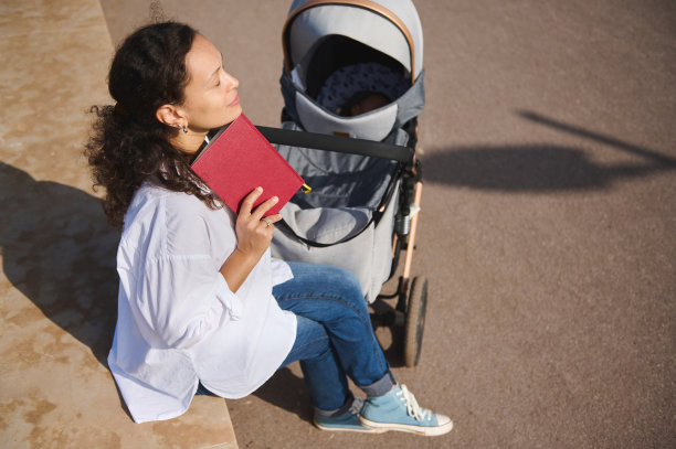 年轻的妈妈喜欢在户外读书，在公园里放松，和她可爱的宝宝一起睡在婴儿车里。图片下载