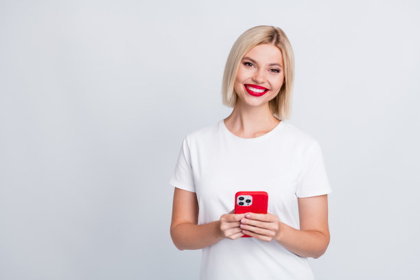 照片中，长着牙齿、笑容灿烂的聪明女孩梳着波波头，穿着白色t恤，在灰色背景下拿着智能手机聊天图片下载