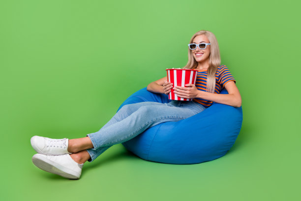 全身尺寸的照片微笑的快乐的女士坐在豆袋拿桶爆米花零食戴3d眼镜孤立的绿色背景图片下载