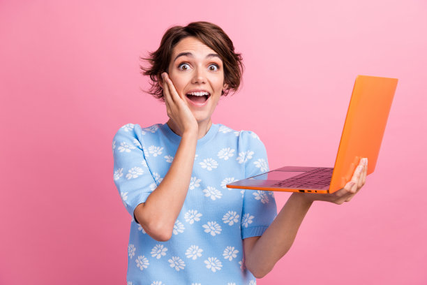 印象深刻的开朗女孩的照片，手触摸脸颊，抱无线笔记本电脑孤立在粉红色的背景图片下载