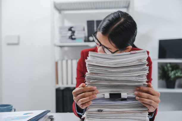 压力很大的人戴着眼镜，穿着红色运动夹克，一只手拿着头，另一只手拿着笔，旁边放着一大堆文件。图片下载