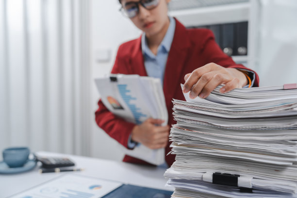 压力很大的人戴着眼镜，穿着红色运动夹克，一只手拿着头，另一只手拿着笔，旁边放着一大堆文件。图片下载