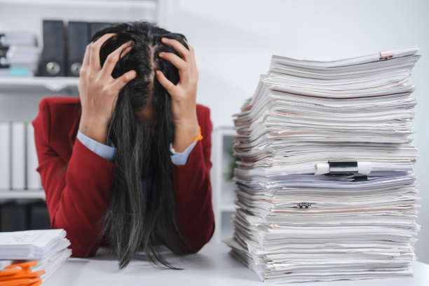 压力很大的人戴着眼镜，穿着红色运动夹克，一只手拿着头，另一只手拿着笔，旁边放着一大堆文件。图片下载