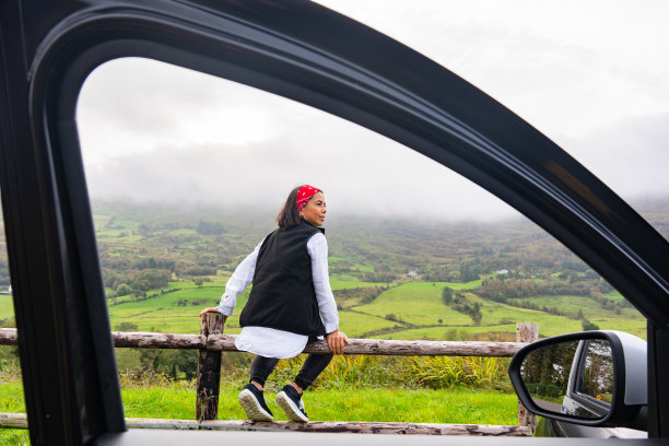 一名女子坐在木栅栏上，欣赏着绿色田野和低云的美丽景色，这张照片是从她租来的汽车的窗户拍摄的图片下载