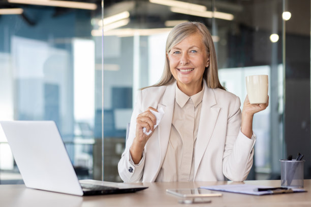 一个面带微笑、头发花白的女商人坐在办公室的桌子旁，拿着杯子和餐巾，对着镜头微笑图片下载
