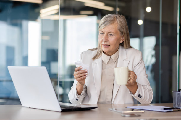 生病的高级商务女性在办公室工作，坐在桌子旁，手里拿着杯子和餐巾，担心地看着笔记本电脑的屏幕图片下载