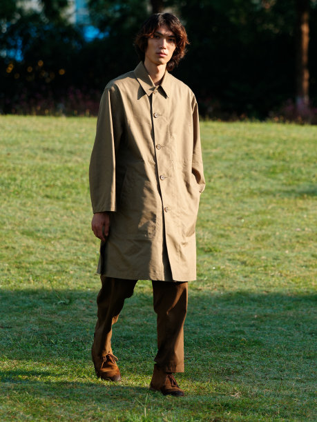 一个英俊的中国年轻人的肖像，他穿着卡其布大衣，一头卷发，在阳光明媚的日子里走在绿色的草地上，男性时尚，凉爽的亚洲年轻人的生活方式。图片下载