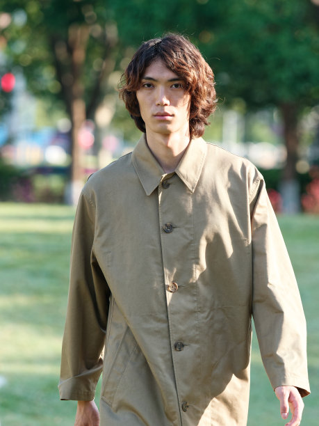 一个英俊的中国年轻人的肖像，他穿着卡其布大衣，一头卷发，在阳光明媚的日子里走在绿色的草地上，男性时尚，凉爽的亚洲年轻人的生活方式。图片下载