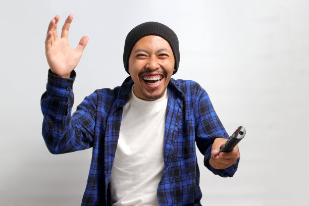 一名亚洲男子手持电视遥控器观看电视上的体育比赛图片下载