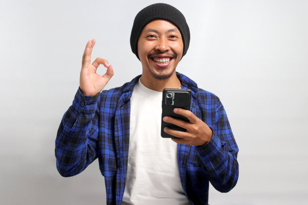 一名亚洲男子拿着智能手机做了一个OK的手势图片下载