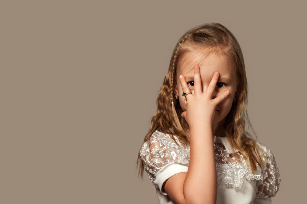 可爱的封面女孩孩子5-6岁，捂着脸，手指上戴着戒指，看着相机。小女孩的肖像在米色孤立的背景下，工作室拍摄。A代概念。复制广告文本空间图片下载