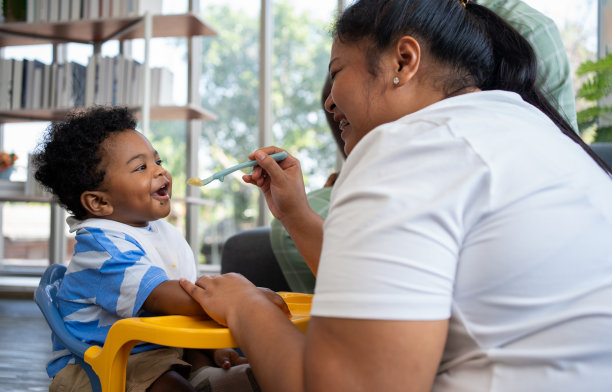 亚洲妈妈在给她9个月大的可爱宝宝喂奶，非裔美国人在家帮忙拿盘子。照片系列的家庭，孩子和快乐的人的概念。父母喂养孩子。图片下载